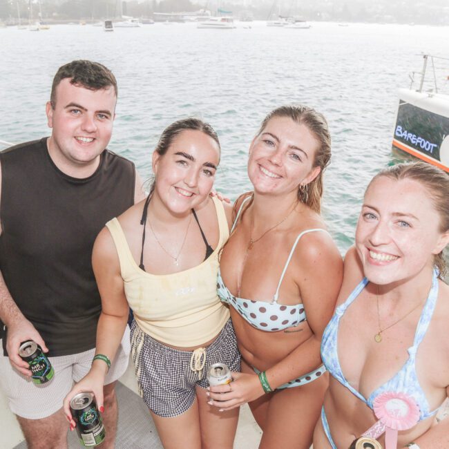 Four smiling young adults in swimwear stand on a boat holding drinks, with water, sailboats, and a catamaran in the background. The scene appears relaxed and joyful, likely during a daytime outing.