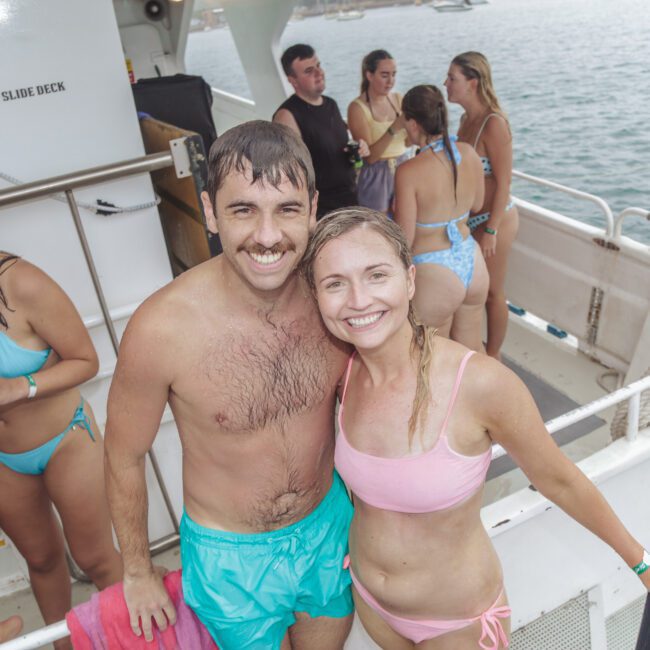A smiling man and woman in swimsuits pose together on a boat deck with other people in swimwear in the background. The group appears to be enjoying a day on the water.