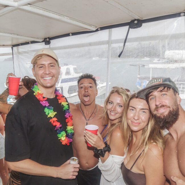 A group of young adults in swimwear and party attire smile and pose together on a boat, holding drinks. The background shows boats on the water and a cloudy sky. The scene looks lively and festive.
