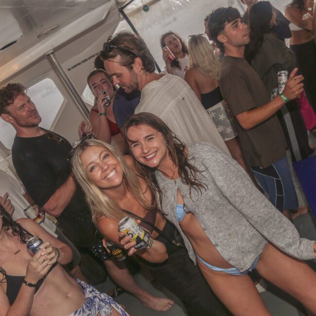A group of young adults in swimsuits and casual clothes are smiling and holding drinks at a lively boat party. Some people are dancing and chatting. The atmosphere is fun and social, with wet hair suggesting recent swimming.