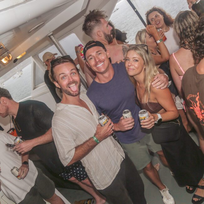 A group of people on a boat at a lively social event. Three people in the center smile and pose for the camera, holding canned drinks, while others chat and socialize in the background.