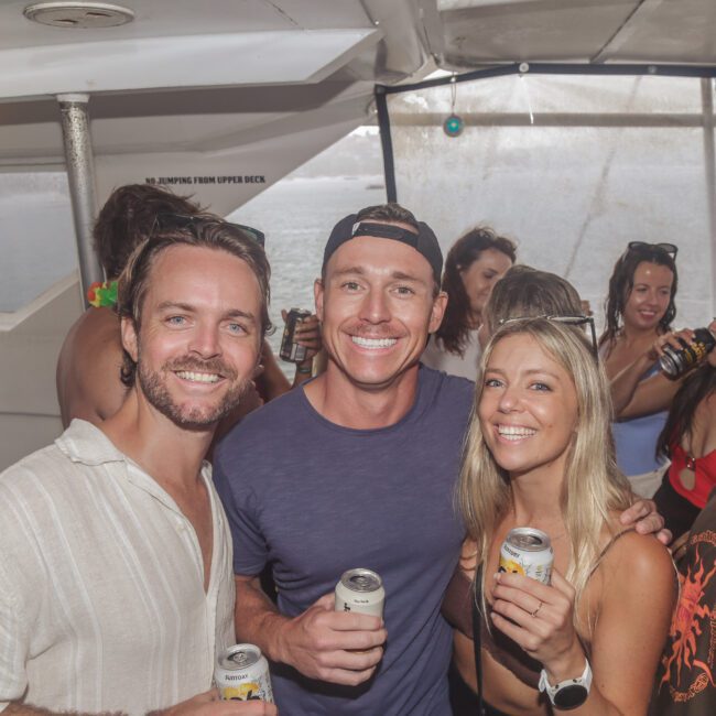 A group of people enjoying a boat party. Three people in front, smiling and holding canned drinks, with others socializing in the background. The setting appears casual and festive inside a boat cabin.