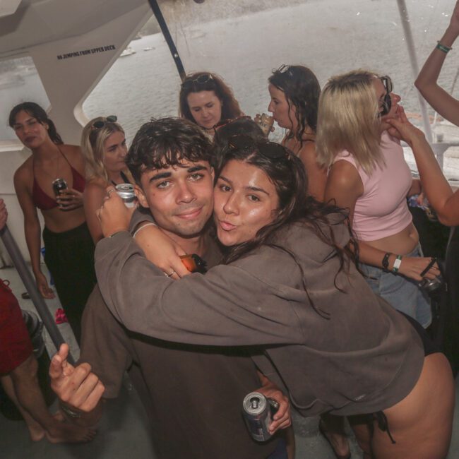 A group of young adults are partying on a boat. Two people in the foreground hug and pose for the camera, both holding drinks, while others socialize and dance in the background. Water is visible outside the boat windows.