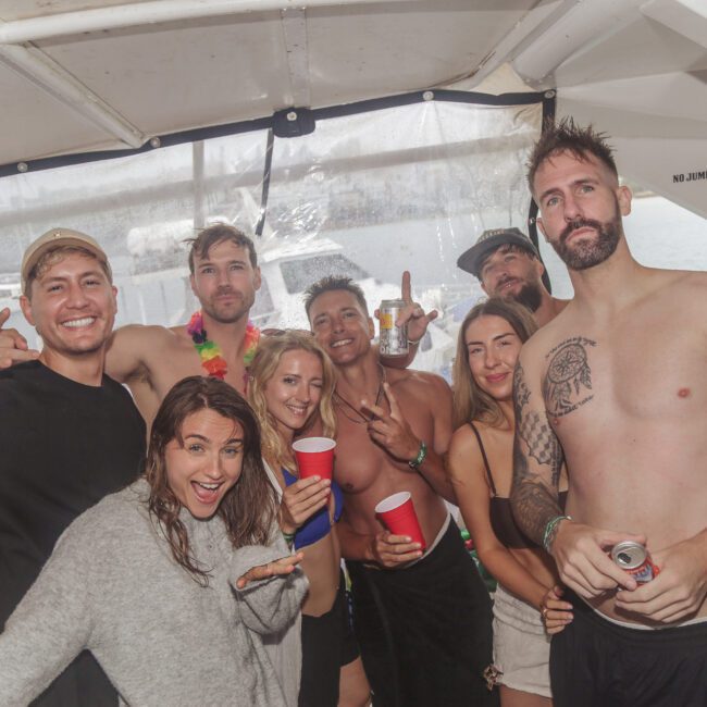 A group of smiling adults in swimsuits and casual clothing pose together on a boat, some holding red cups. The atmosphere is festive and relaxed, with the ocean visible through the windows behind them.
