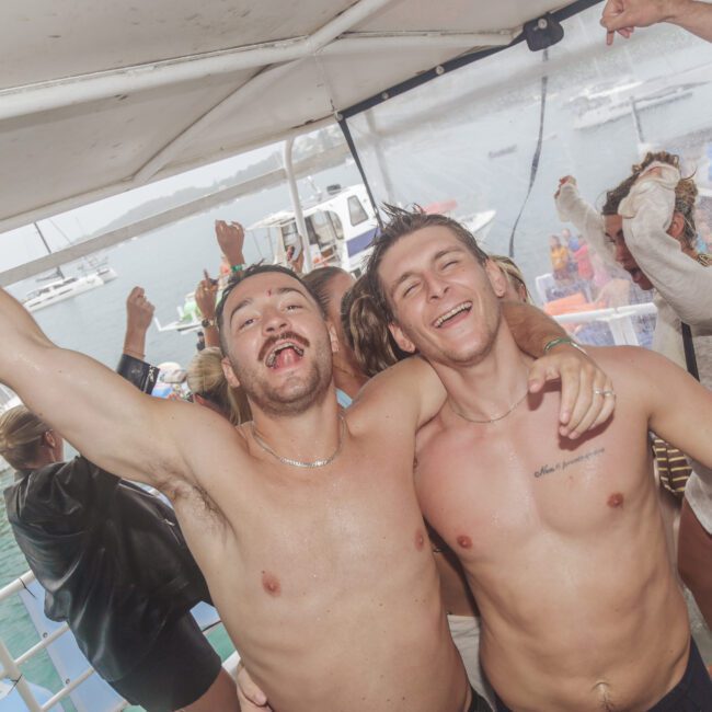 Two young men with bare chests smile and pose with arms around each other on a boat, while others dance and celebrate in the background near water and other boats.