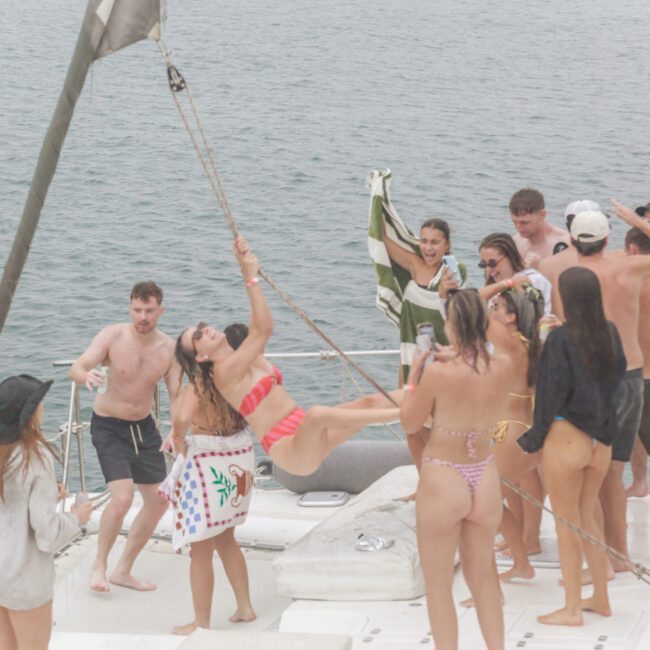 A group of people in swimsuits dance and have fun on a boat. One woman swings on a rope, while others laugh, dance, and hold drinks. The boat is on the water, and the atmosphere appears lively and festive.