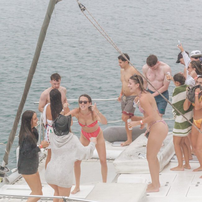 A group of people in swimsuits laugh, dance, and enjoy drinks together on the deck of a sailboat at sea on a cloudy day.