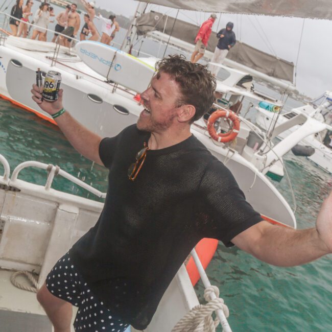 A man in wet clothes, holding a drink and pointing, dances joyfully on a boat with other people and catamarans in the background on the water. The mood is lively and festive despite overcast weather.