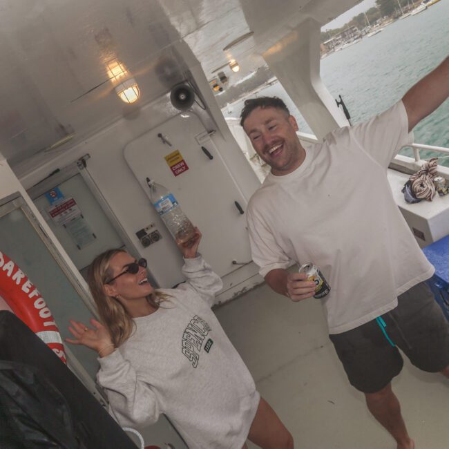 Two people stand and dance on the deck of a boat, smiling and holding drinks. The woman wears sunglasses and a sweatshirt, while the man wears a white T-shirt and shorts. Water and shoreline are visible in the background.
