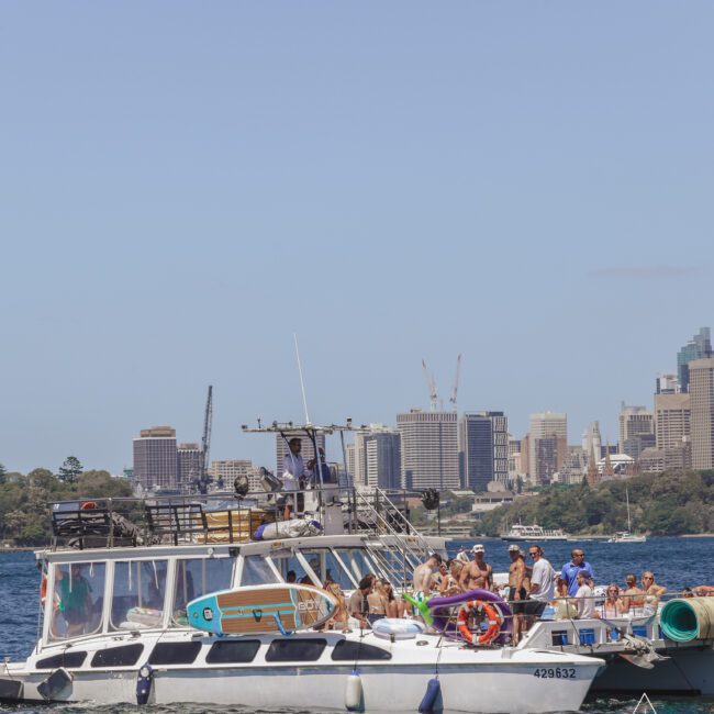A group of people enjoying a sunny day on a white yacht in the harbor, with city buildings and cranes visible in the background under a clear sky.