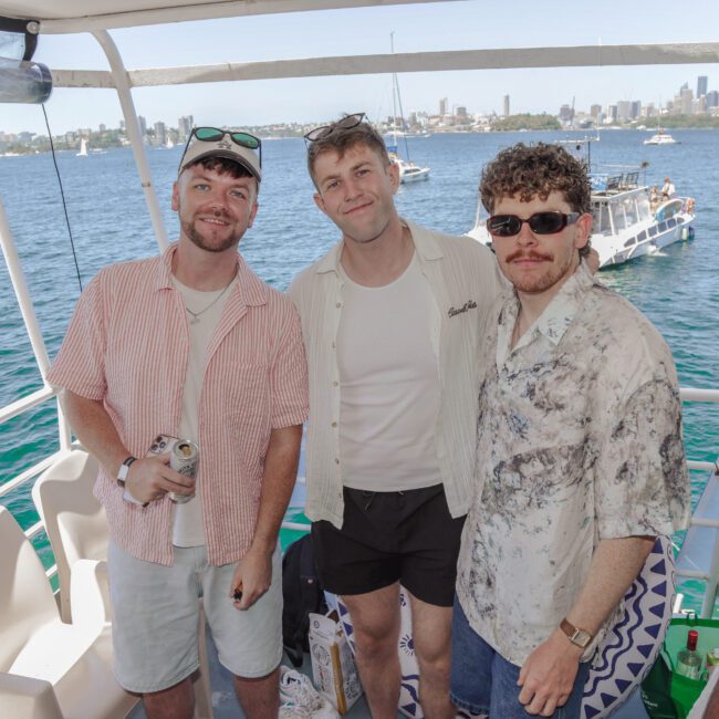 Three men pose and smile on a boat with water and other boats in the background; they are wearing sunglasses and casual summer clothes on a sunny day, with a city skyline visible in the distance.