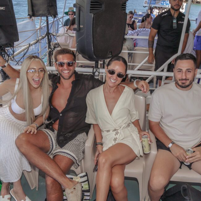 Four adults sit together on a boat, smiling and holding drinks. Two women wear sunglasses and light outfits; two men sit beside them, one smiling in a black shirt, the other in a beige shirt. The sea is visible in the background.