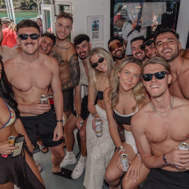 A group of young adults in swimwear, smiling and posing together on a boat with drinks in hand. The background shows blue water and greenery, suggesting a fun, sunny day out.