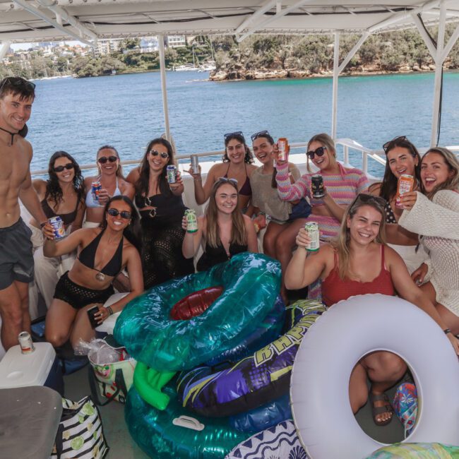 A group of smiling people sit together on a boat, holding up drinks and surrounded by colorful pool floats, with water and greenery in the background on a sunny day.