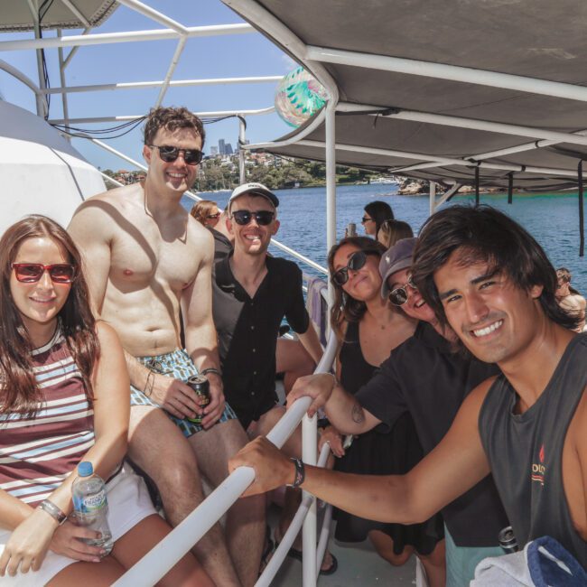 A group of six young adults, smiling and posing together on the deck of a boat under sunny weather, with blue water and a shoreline in the background. Some are wearing sunglasses and summer clothes.