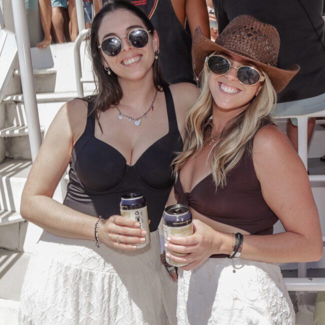 Two women smiling and holding canned drinks at an outdoor event. Both wear sunglasses, white skirts, and dark tops; one also wears a brown cowboy hat. They stand close together in a sunny, casual setting with people in the background.
