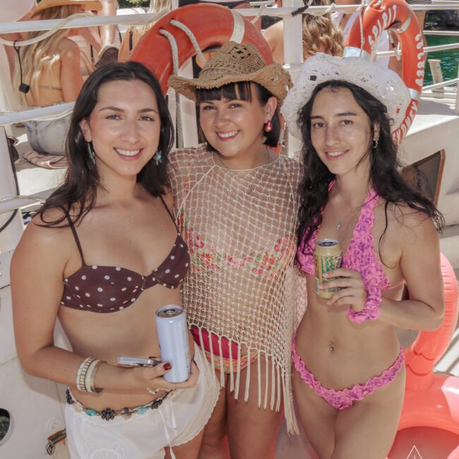 Three women in swimsuits and hats smile while holding drinks on a boat during a sunny day, with other people and life preservers in the background.