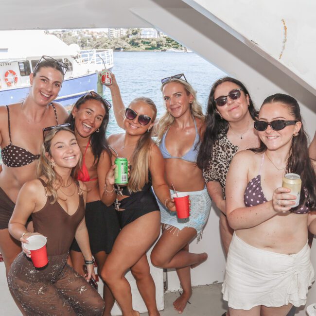 Seven women in swimsuits smile and pose together on a boat, holding drinks. The background shows water, a blue and white boat, and some shoreline with buildings. Sunlight brightens the lively scene.