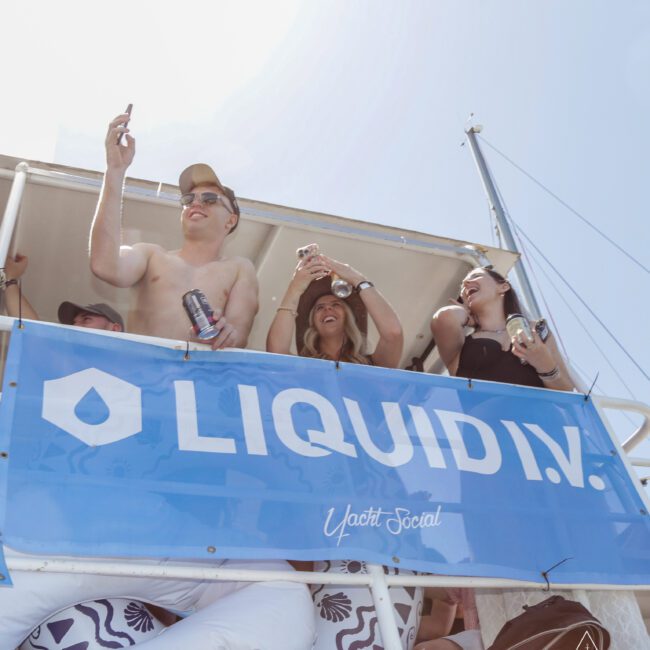 A group of people in swimwear stand on a boat deck behind a large blue "LIQUID I.V." banner, smiling and holding drinks, enjoying a sunny day. The sky is clear and bright above them.