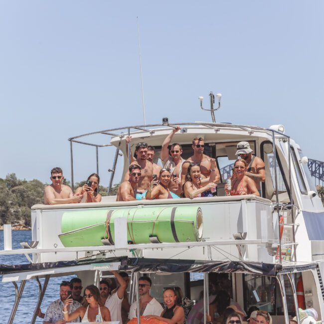 A group of people in swimsuits stand and sit on the upper deck of a boat, smiling and posing for a photo on a sunny day. The city skyline and a bridge are visible in the background across the water.