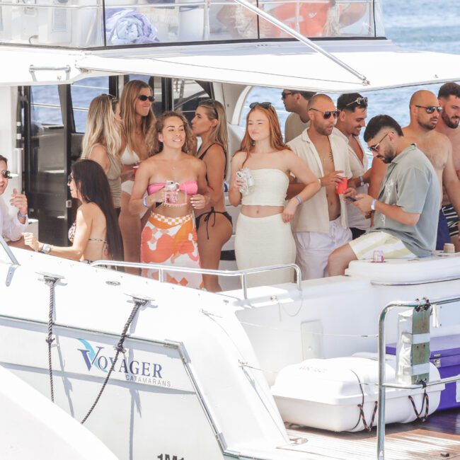 A group of people in swimwear enjoy drinks and conversation on a white yacht named Voyager, docked by the water on a sunny day. Coolers and bags are visible onboard.