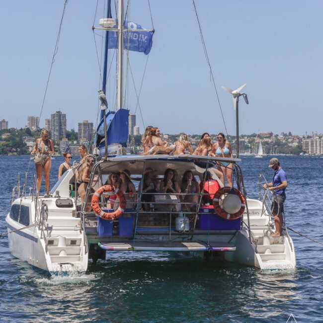 A group of people relax and socialize on the deck of a catamaran sailing on a sunny day, with a city skyline and waterfront buildings visible in the background.
