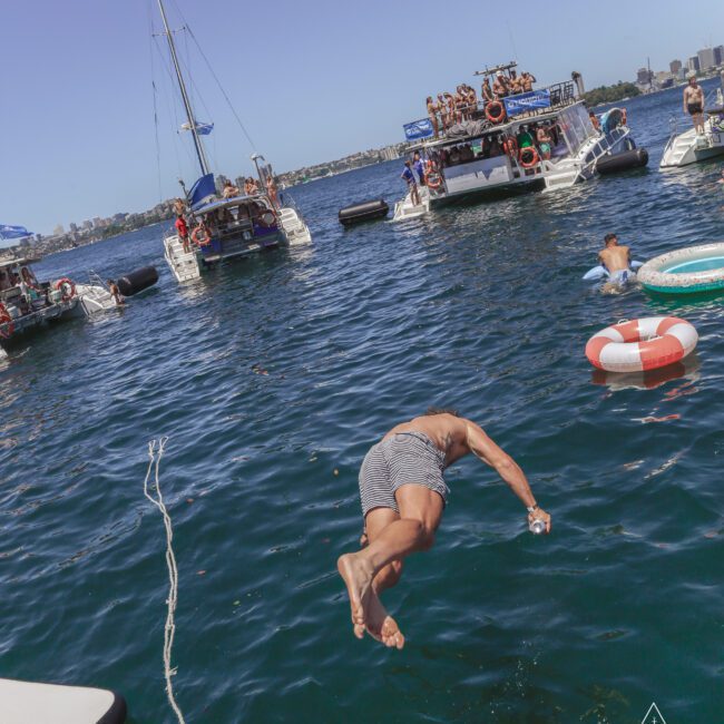 A man in swim trunks dives into a lake from a boat, surrounded by several party boats with people, while others swim and float nearby on a sunny day.
