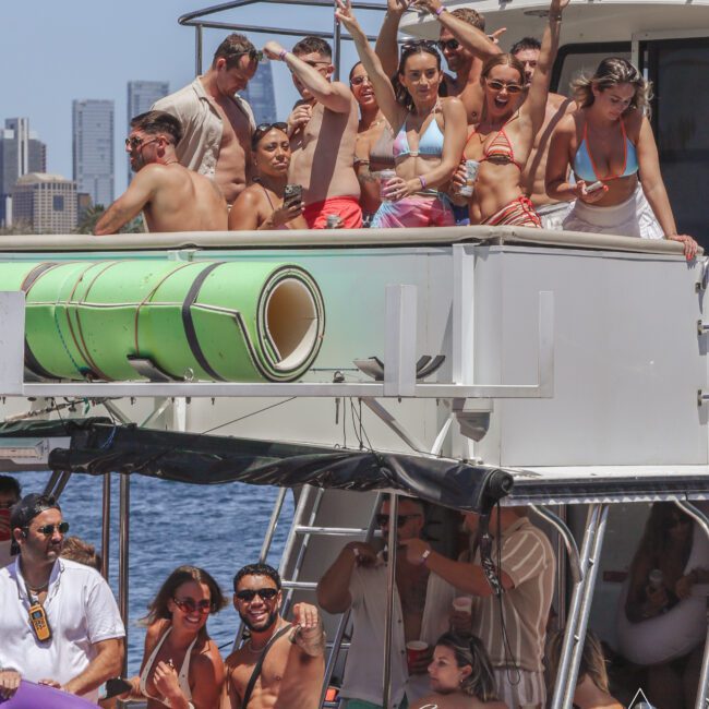 A group of people in swimsuits are celebrating and posing on a boat deck, some with arms raised. The boat has rolled-up green mats on the roof, and a city skyline is visible in the background under a clear sky.
