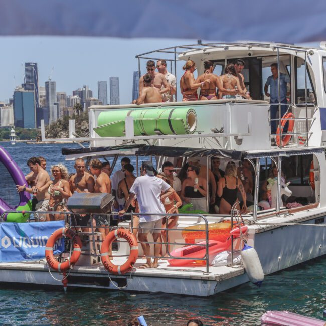A group of people in swimsuits enjoy a sunny day on a boat, chatting and relaxing. The boat is on the water near a city skyline with tall buildings and the Sydney Harbour Bridge in the background.