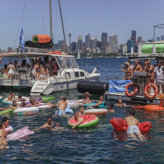 People relax on boats and colorful pool floats in the water near a city skyline on a sunny day. The scene is lively and festive, with groups socializing and swimming. The city buildings are visible in the background.