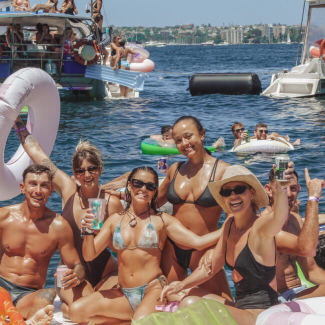A group of smiling people in swimsuits pose on a boat, holding drinks and inflatable floaties, with other people and boats on the water in the sunny background.