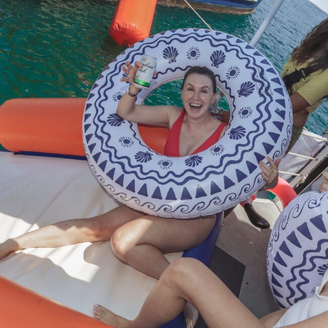 A smiling woman in a red swimsuit sits in a patterned pool float, holding up a can, surrounded by friends on a boat near the ocean. The mood is cheerful and fun.
