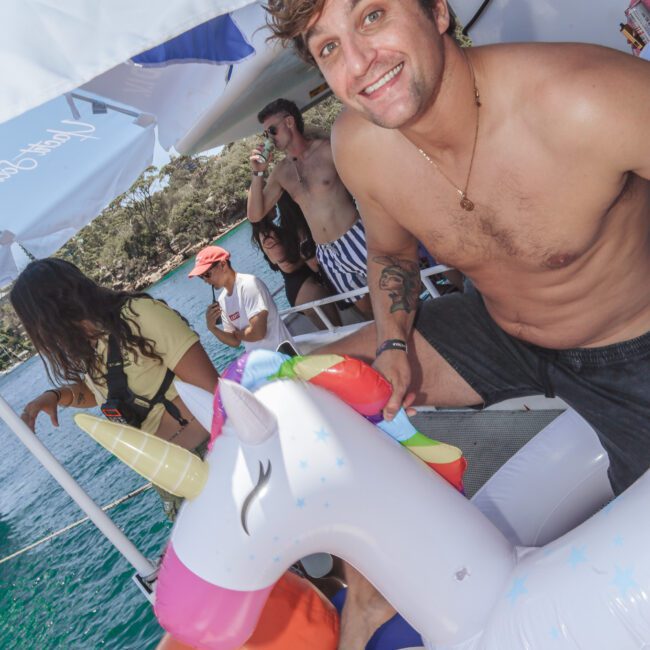 A smiling shirtless man holds a large inflatable unicorn on a boat with several people relaxing around him; water and trees are visible in the background under sunny skies.