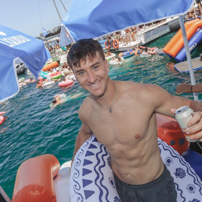A smiling young man in swim trunks holds a can while standing on a float at a lively pool party, with many people and floats in the water. There are umbrellas, boats, and a festive atmosphere.