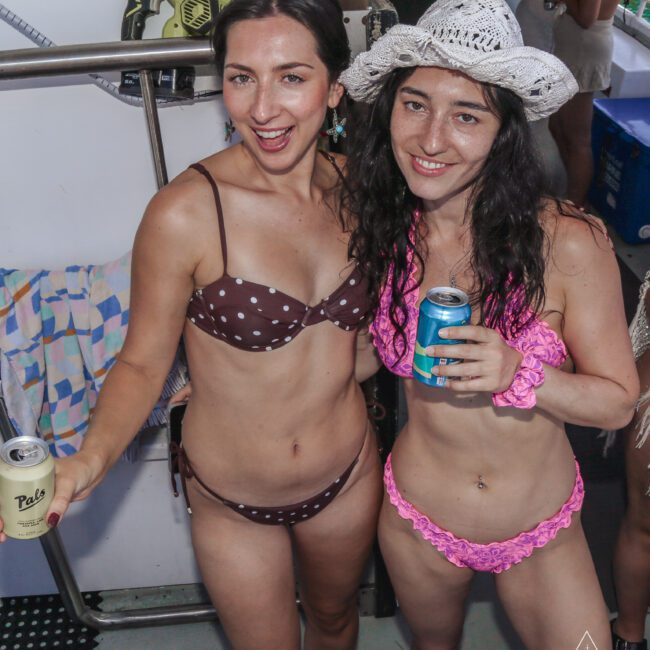 Two women in swimsuits pose and smile at a party. One wears a brown polka dot bikini and holds a drink; the other wears a pink ruffle bikini, a white cowboy hat, and holds a can. They stand by a rail with drinks in hand.