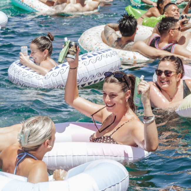 A group of people relax and smile on inflatable pool floats in clear blue water, holding drinks and enjoying a sunny day. The scene is lively, social, and festive, with everyone appearing to have fun.
