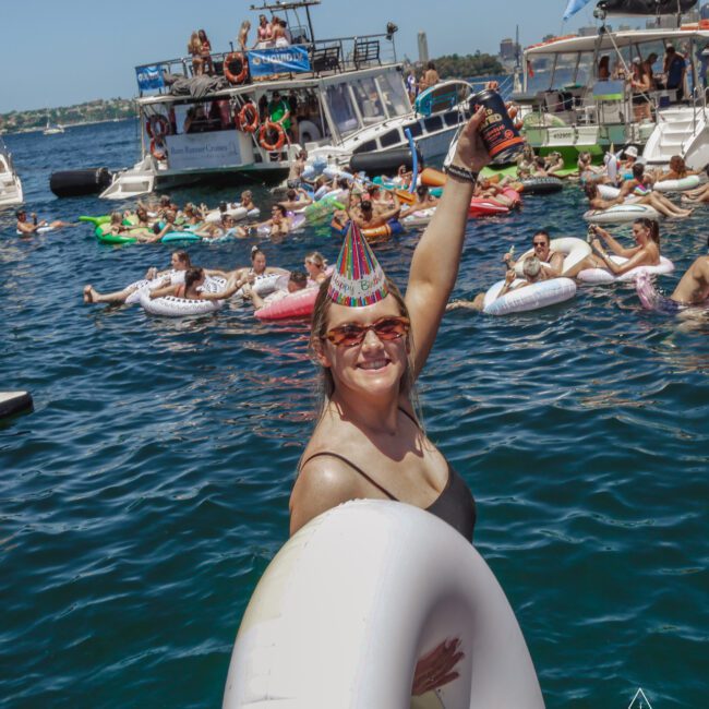 A woman wearing a party hat smiles at the camera while holding a drink and sitting on an inflatable ring. She is surrounded by people on floats in the water and several boats during a lively outdoor party.