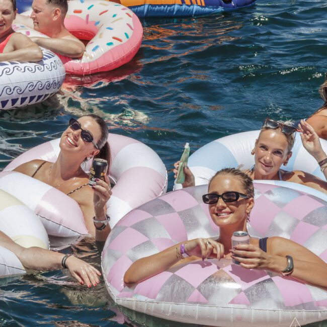 People relaxing in colorful donut-shaped pool floats on blue water, smiling and holding drinks, wearing sunglasses and swimsuits. The atmosphere is cheerful and summery.