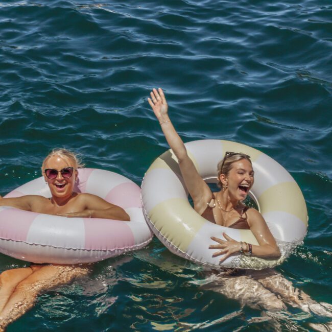 Two women float on colorful inflatable rings in deep blue water, smiling and waving. They wear sunglasses and swimsuits, enjoying a sunny day. The Yacht Social logo is visible in the lower right corner.
