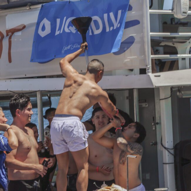 A group of young adults in swimwear enjoy a party on a boat. One person pours a drink from a funnel into another’s mouth, while others look on and smile. A blue “Liquid I.V.” banner hangs in the background.