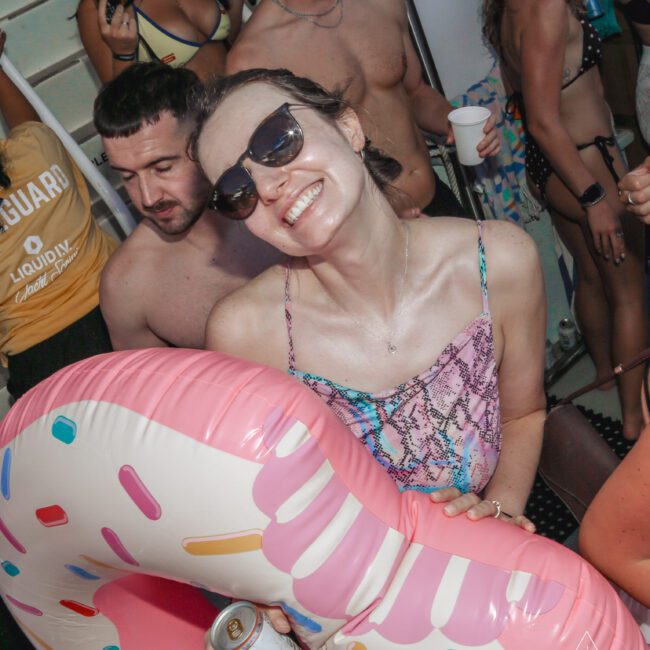 A woman in sunglasses smiles at the camera while holding a pink donut-shaped pool float at a lively pool party. People in swimwear are around her, some holding drinks, with a festive summer atmosphere.