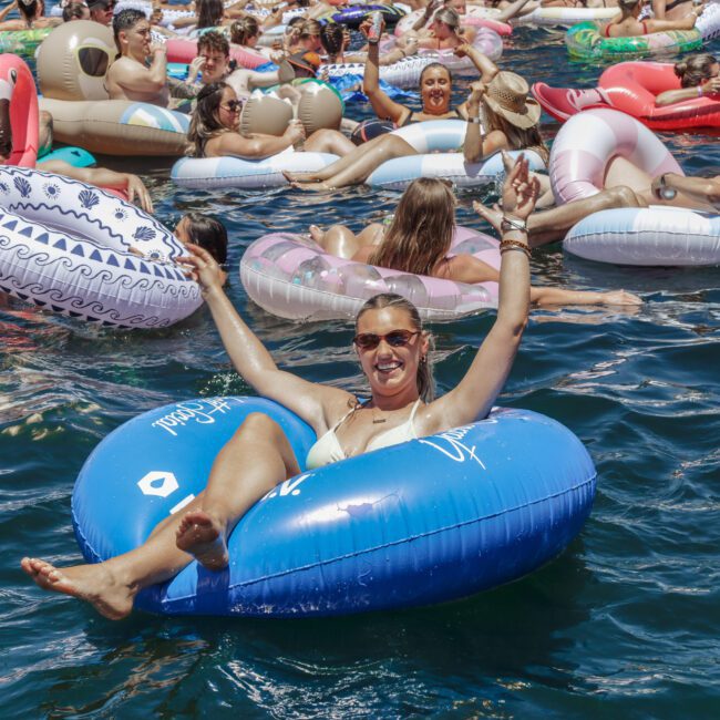 A smiling woman in sunglasses relaxes on a blue inflatable tube in a crowded pool, surrounded by people on colorful floaties, enjoying a sunny day.