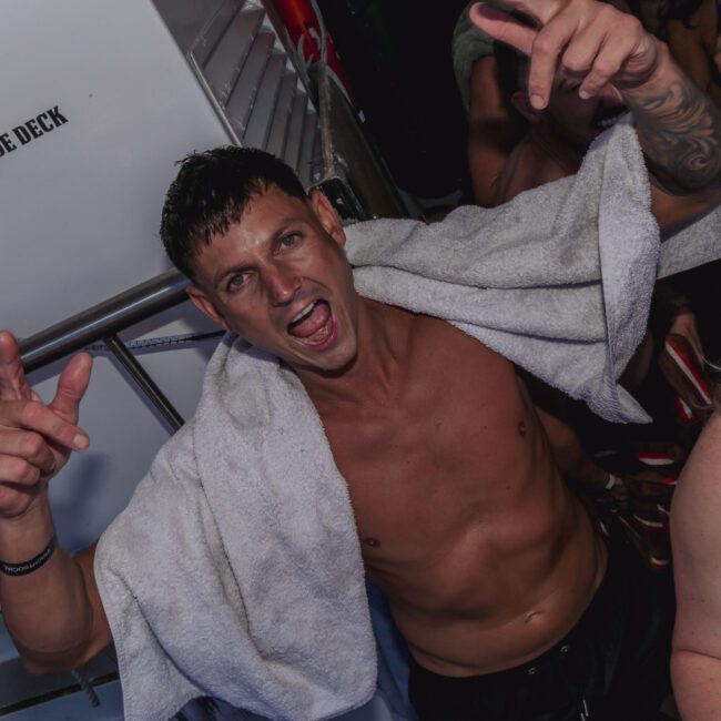A man with a towel draped over his shoulders stands on a boat, making hand gestures and smiling energetically at a party. Other people in swimwear are around him, and a sign reads "NO PERSONS ON SLIDE DECK.