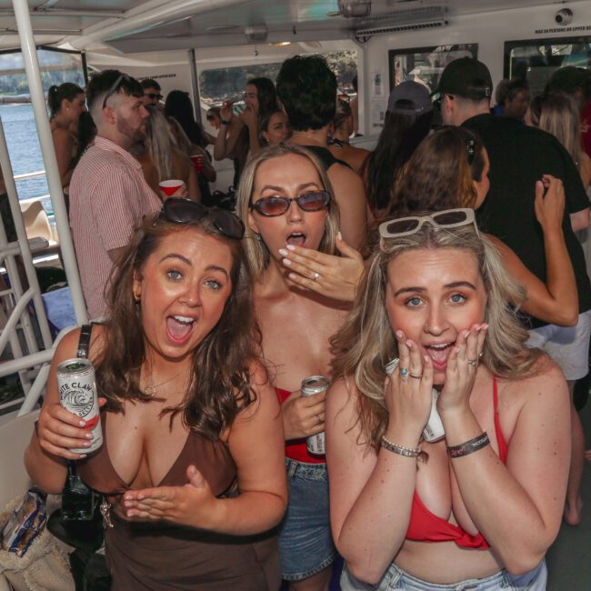 Three women in swimsuits pose playfully with surprised expressions on a crowded party boat, holding drinks, while other people mingle in the background. Sunlight streams in from the side, and water is visible outside.