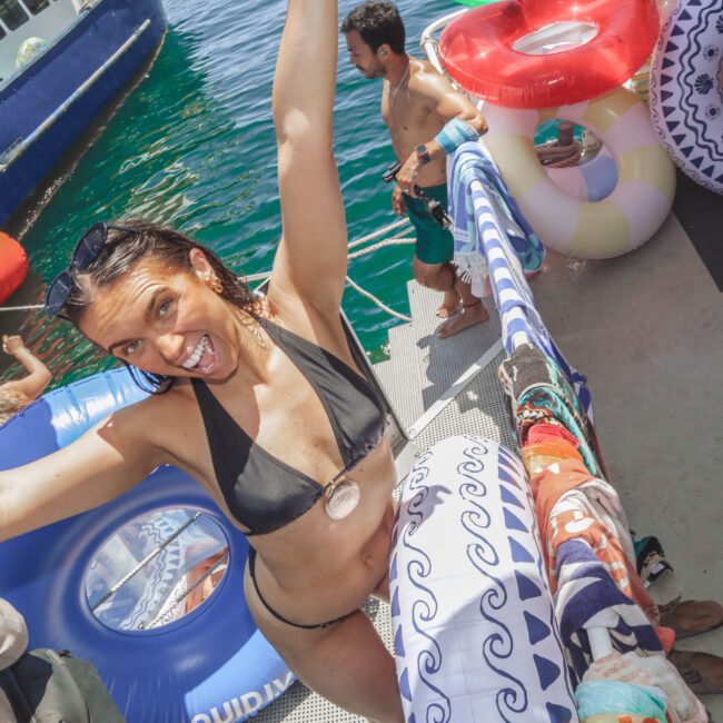 A woman in a black bikini smiles and raises her arms in excitement on a dock by the water, surrounded by colorful pool floats and towels, with other people and a boat nearby.
