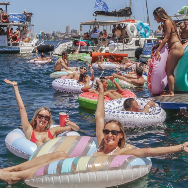 People relaxing on colorful inflatable pool floats in the water near docked yachts, enjoying a sunny day. Many are smiling, raising their arms, and wearing sunglasses, with boats and blue sky in the background.