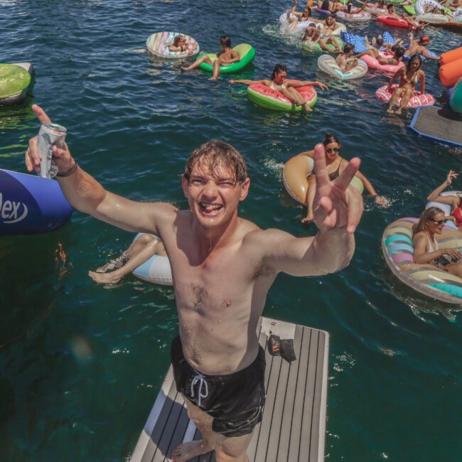 A smiling young man in swim trunks stands on a dock holding a drink and making a peace sign, with people relaxing on colorful pool floats in the water behind him. The scene is lively and sunny, with a party atmosphere.
