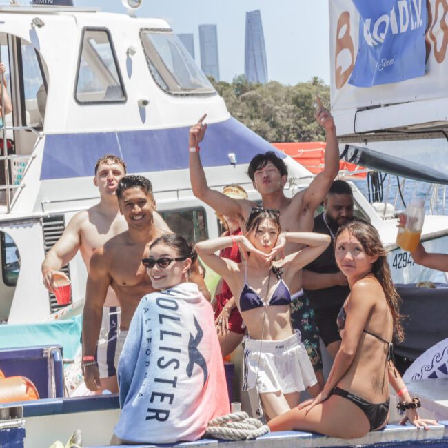 A group of young people in swimwear celebrate on a boat, some holding drinks and smiling at the camera. Other boats and buildings are visible in the background on a sunny day.