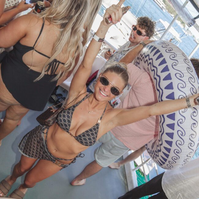 A woman in a patterned bikini, sunglasses, and beach hat smiles with arms raised at a lively boat party. People around her are chatting and holding drinks, with water and boats visible in the background.