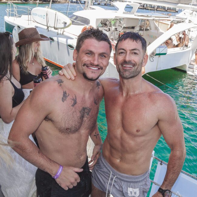 Two smiling shirtless men stand side by side on a boat, posing for a photo. Other people and a white yacht are visible in the background on a sunny day by the water.